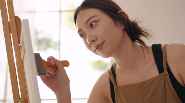 Creative young Asian woman artist painting on canvas with brush in bright home studio. Relaxed female hobbyist enjoying peaceful indoor leisure time, expressing imagination for art.
