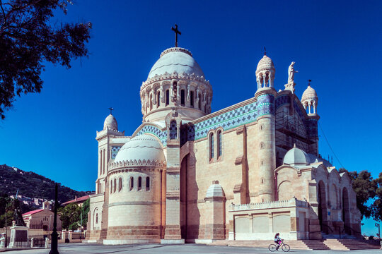 View of the Basilica of Our Lady of Africa with its distinctive neo-Byzantine architecture, stone walls, and a person cycling in the foreground in Algiers, Algeria.