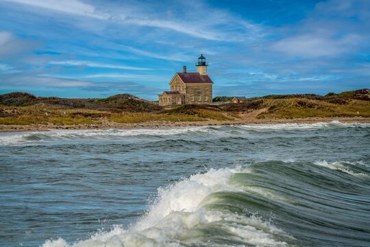 View of the historic Block Island North Light on grassy dunes above the beach with crashing waves in the foreground under a blue sky in New Shoreham, Rhode Island, United States.