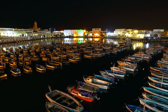 View of numerous small fishing boats moored in a calm harbor at night with illuminated buildings along the waterfront in Algeria.