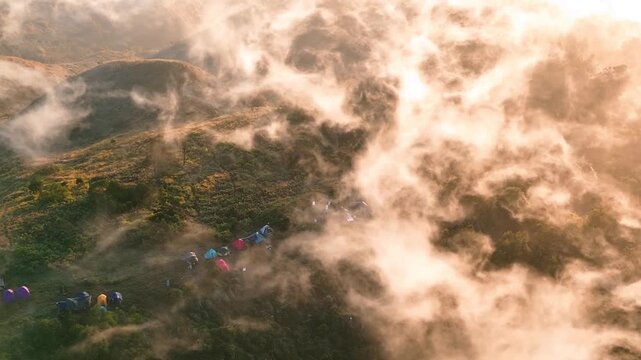 Aerial view of colorful tents pitched on the slopes of Gunung Prau surrounded by thick morning mist and golden sunlight in Wonosobo, Jawa Tengah, Indonesia.