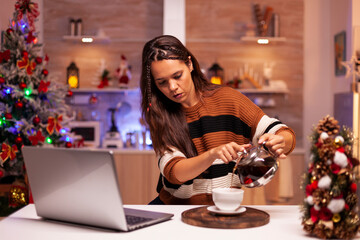 Festive woman fills a mug from a kettle while using a laptop to exchange Christmas greetings with friends at home. © DC Studio