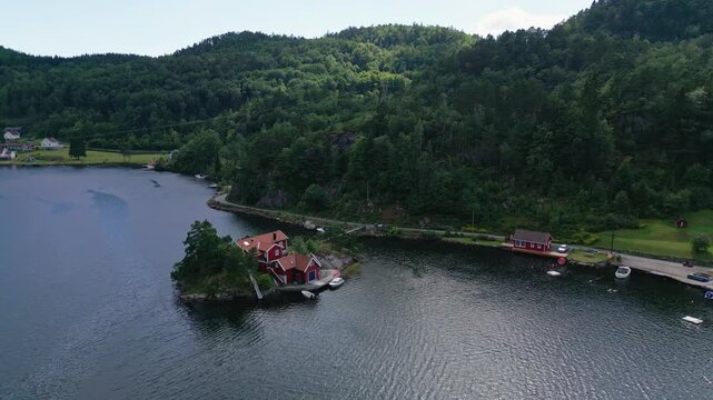 Aerial view of Sogne featuring traditional red wooden houses along the shoreline with boats and lush green hills in Kristiansand, Agder, Norway.