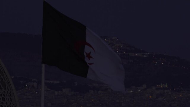 Aerial view of the Algerian flag waving against a backdrop of illuminated city buildings at night in Algeria.