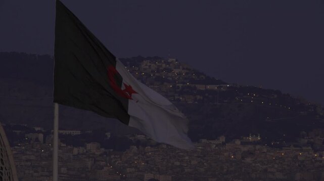 Aerial view of the Algerian flag waving in the wind against a backdrop of a hillside city at dusk in Algeria.