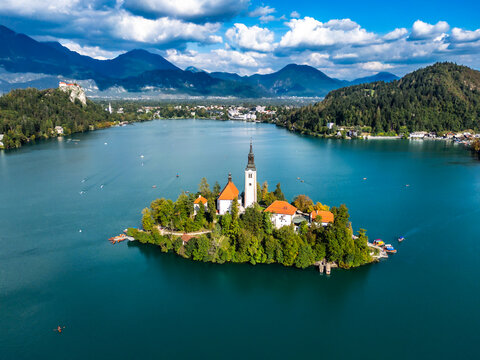 Aerial view of Lake Bled with its iconic island church surrounded by turquoise water and lush green forests under a blue sky with clouds in Bled, Radovljica, Slovenia.
