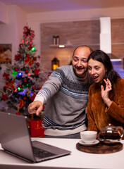 Couple talks with remote friends through a video call, sharing Christmas greetings from their decorated kitchen at home. © DC Studio