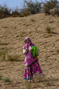 Nagarparkar, Sindh, Pakistan - 08 January 2015: View of a woman in a traditional pink dress carrying a green bag while walking across the dry, sandy terrain of the Thar Desert.