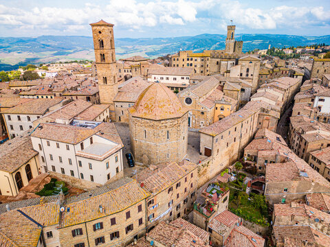 Aerial view of the historic Baptistery of San Giovanni and Cathedral of Santa Maria Assunta surrounded by terracotta rooftops and stone architecture in Volterra, Tuscany, Italy.