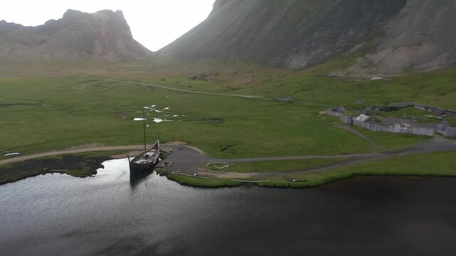 Aerial view of a Viking ship replica moored on the shore of a calm pond with green grassy fields and dramatic mountains in the background at Stokksnes, Myrdalshreppur, Iceland.