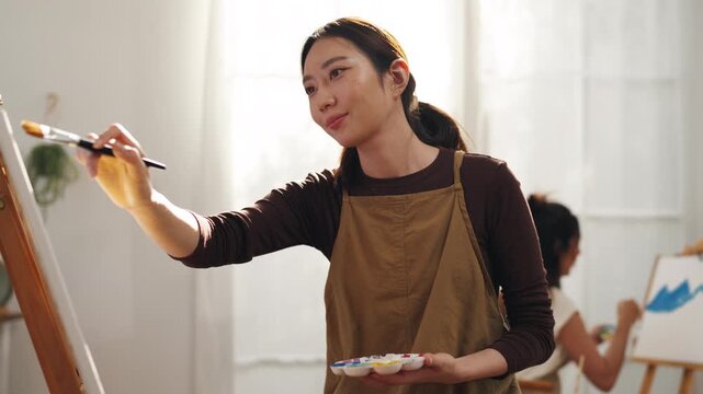 Creative young Asian woman artist painting on canvas with brush in bright home studio. Relaxed female hobbyist enjoying peaceful indoor leisure time, expressing imagination for art.