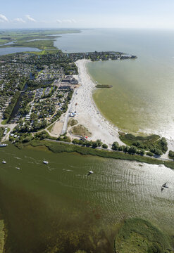 Aerial view of De Holle Poarte beach along the IJsselmeer coast with holiday park facilities, sandy shoreline, and sailboats on the water in Makkum, Friesland, Netherlands.