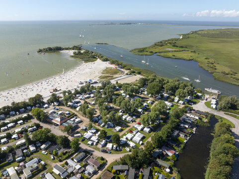 Aerial view of De Holle Poarte beach and campsite with numerous caravans, lush trees, and boats on the water in Makkum, Friesland, Netherlands.