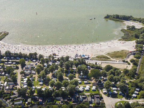 Aerial view of De Holle Poarte beach and adjacent campsite with numerous caravans, lush trees, and people enjoying the sandy shore and water in Makkum, Friesland, Netherlands.