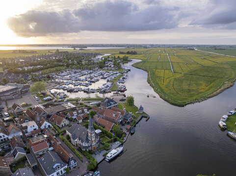 Aerial view of the historic village of Woudsend with its marina, canal, and church surrounded by lush green fields under a golden sunset sky in Woudsend, Friesland, Netherlands.