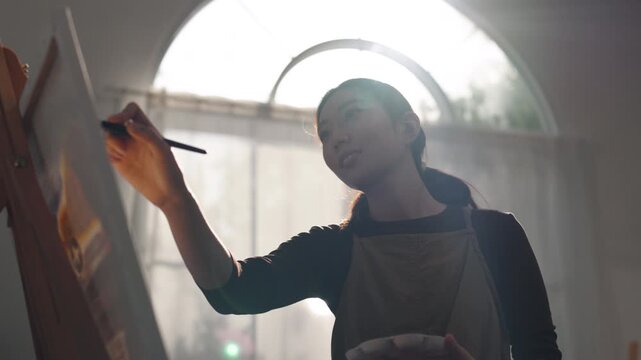 Creative young Asian woman artist painting on canvas with brush in bright home studio. Relaxed female hobbyist enjoying peaceful indoor leisure time, expressing imagination for art.