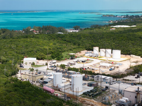 Aerial view of a fuel storage facility with large white storage tanks, industrial buildings, and tanker trucks near the turquoise waters of George Town, Exuma, The Bahamas.