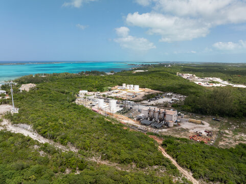 Aerial view of an industrial power plant facility with fuel and storage tanks surrounded by dense tropical vegetation near the turquoise coastline of George Town, Exuma, The Bahamas.
