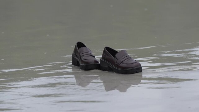 Loafer shoe footwear pair of dark leather slip on on wet pavement puddle after rain, reflection on soaked street, beach shoreline sand tidal pool, abandoned floating footwear on calm water