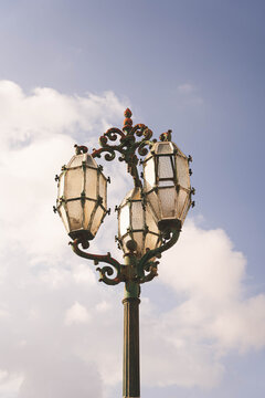 Valletta, Port Region, Malta - 14 July 2022: View of an ornate vintage street lamp with three glass lanterns against a blue sky with soft clouds.