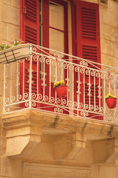 Valletta, Malta - 14 July 2022: View of a traditional Maltese balcony with red shutters, white wrought iron railing, and hanging pots with yellow flowers against a limestone wall.