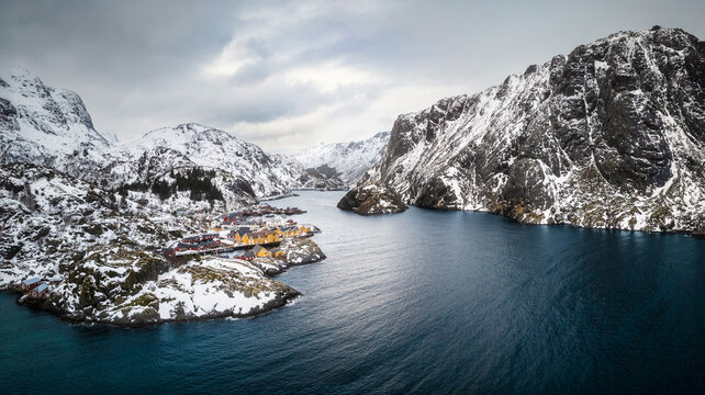 Aerial view of Lofoten featuring traditional cabins nestled along the rocky coastline, surrounded by snow-covered mountains and deep blue arctic waters in Lofoten, Nordland, Norway.