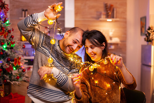 Playful couple becomes caught in glowing tree lights while decorating their Christmas kitchen and laughing together.
