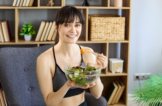 Woman eating healthy salad at home. Smiling fitness woman holds a bowl of greens and tomatoes with a wooden fork. Ideal for diet, nutrition and wellness campaigns. Pure healthy living vibe now.