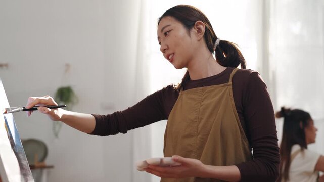 Creative young Asian woman artist painting on canvas with brush in bright home studio. Relaxed female hobbyist enjoying peaceful indoor leisure time, expressing imagination for art.