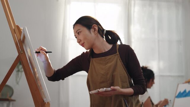 Creative young Asian woman artist painting on canvas with brush in bright home studio. Relaxed female hobbyist enjoying peaceful indoor leisure time, expressing imagination for art.