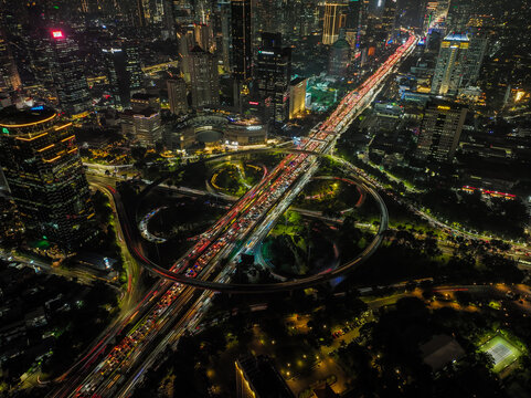 Aerial view of the illuminated Semanggi Bridge at night with heavy traffic light trails on the cloverleaf interchange, surrounded by skyscrapers in Tanah Abang, Jakarta, Indonesia.