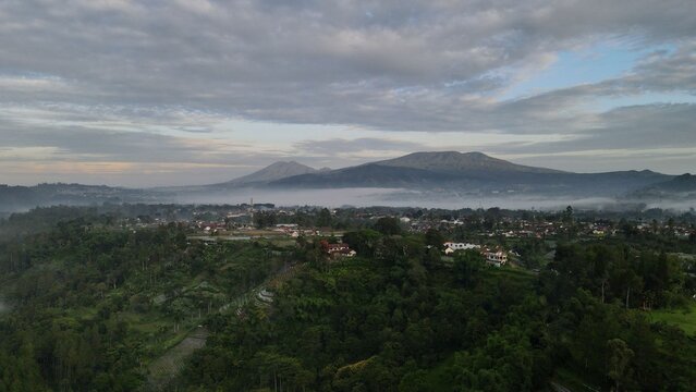 Aerial view of the mountainous landscape with lush green hills, mist, and scattered buildings under a cloudy sky in Lembang, West Java, Indonesia.