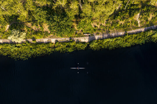 Aerial view of a lone rower in a sculling boat on the dark waters of the Kanal Zeranski, bordered by a lush green tree-lined bank in Warsaw, Masovian Voivodeship, Poland.