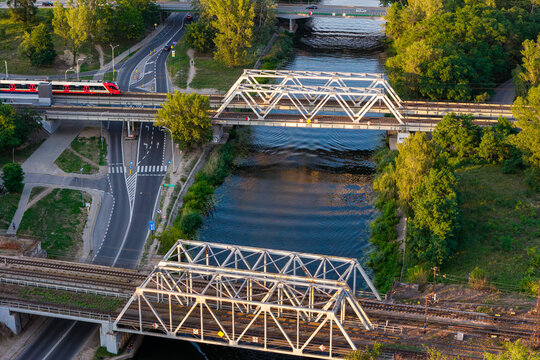 Aerial view of the Kanal Zeranski canal with railway bridges and a train crossing over the water, surrounded by lush green trees and urban roads in Warsaw, Masovian Voivodeship, Poland.