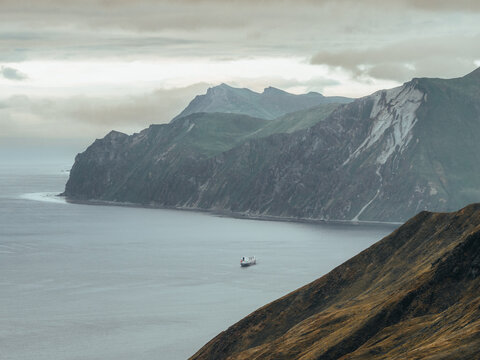 Aerial view of a ship sailing through the calm waters of the bay, surrounded by rugged mountains and steep coastal cliffs under a cloudy sky in Unalaska, Alaska, United States.