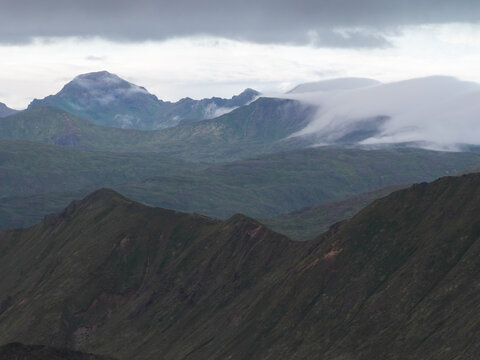 Aerial view of the rugged green mountains of Unalaska shrouded in low-hanging clouds and mist under an overcast sky in Unalaska, Alaska, United States.