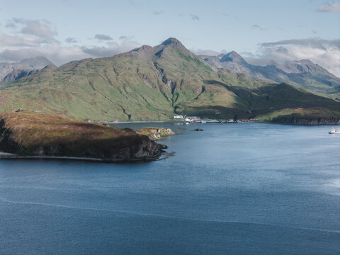 Aerial view of the coastal landscape of Unalaska with green mountains, calm blue waters, and a small settlement along the shoreline in Unalaska, Alaska, United States.