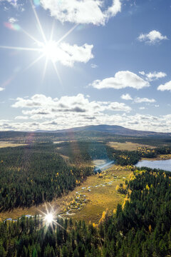 Aerial view of Perilajarvi surrounded by dense coniferous forests, a winding stream, and a lake under a bright sun in Enontekio, Lappi, Finland.