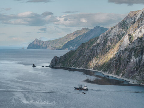 Aerial view of a fishing vessel navigating the coastal waters near the rugged, steep cliffs of Unalaska, Alaska, United States.