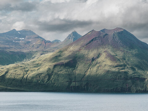 Aerial view of the volcanic mountains of Unalaska with green slopes and reddish rock formations rising above the water under a cloudy sky in Unalaska, Alaska, United States.