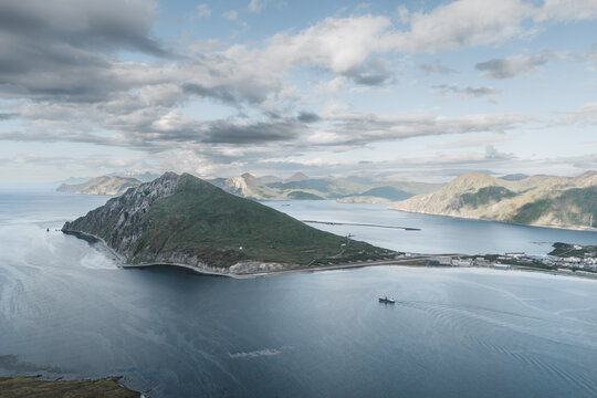 Aerial view of the mountainous landscape of Unalaska surrounded by the Bering Sea with a ship navigating the waters under a cloudy sky in Unalaska, Alaska, United States.