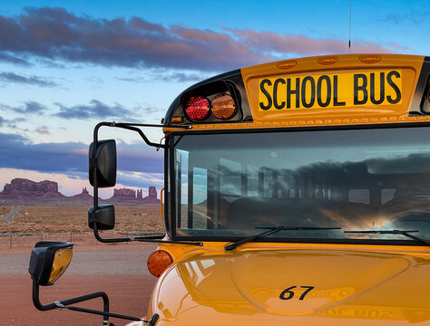 View of a yellow school bus parked in front of the iconic sandstone buttes of Monument Valley Tribal Park under a dramatic sunset sky in Oljato-Monument Valley, Utah, United States.