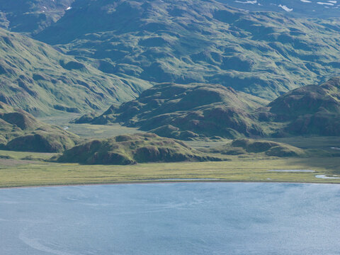 Aerial view of the rugged, green mountainous landscape of Unalaska, featuring rolling hills, coastal waters, and patches of snow on the peaks in Unalaska, Alaska, United States.
