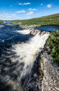 Aerial view of Pikefossen falls with rushing water cascading over rocky ledges surrounded by lush green vegetation under a clear blue sky in Kautokeino, Finnmark, Norway.