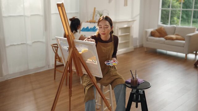 Creative young Asian woman artist painting on canvas with brush in bright home studio. Relaxed female hobbyist enjoying peaceful indoor leisure time, expressing imagination for art.