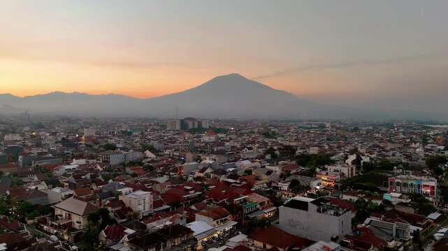 Aerial view of Gunung Arjuno rising above the urban landscape of Malang at sunset, with dense rooftops and warm light in Malang, Jawa Timur, Indonesia.