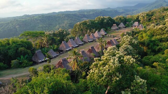 Aerial view of Kampung Adat Belaraghi, a traditional village with thatched-roof houses nestled in lush hills, surrounded by forest in Kecamatan Aimere, Nusa Tenggara Timur, Indonesia.