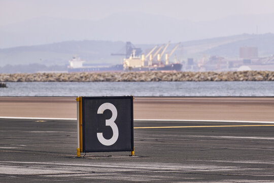 Gibraltar, Gibraltar - 01 March 2026: View of a runway sign with the number 3 at Gibraltar International Airport, with a cargo ship on the water in the background.
