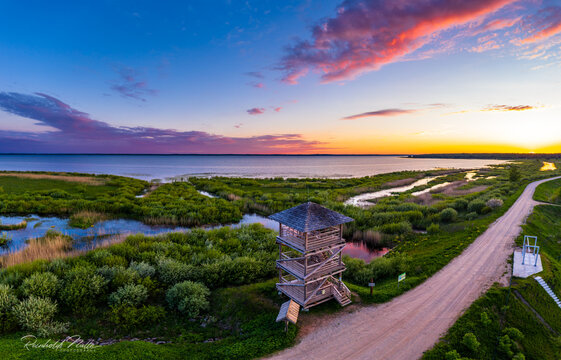 Aerial view of a wooden observation tower situated in a lush wetland area with winding waterways and a dirt road along the shore of a large lake under a vibrant sunset sky in Latvia.
