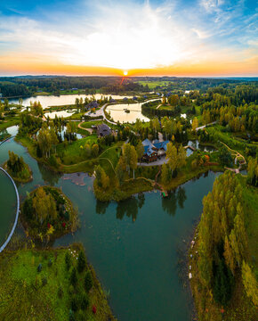 Aerial view of a serene landscape featuring interconnected lakes, lush green forests, and wooden buildings during a golden sunset in Latvia.